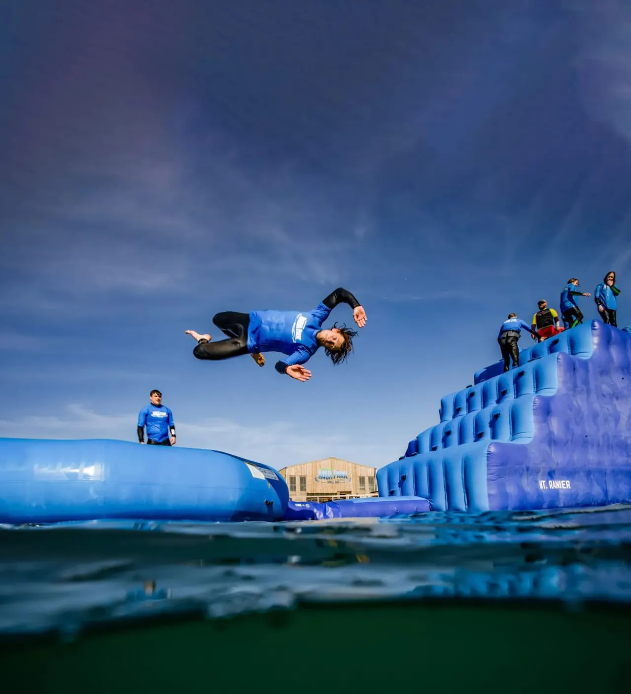 Person in blue launching off an inflatable obstacle course into the sea under a clear sky.