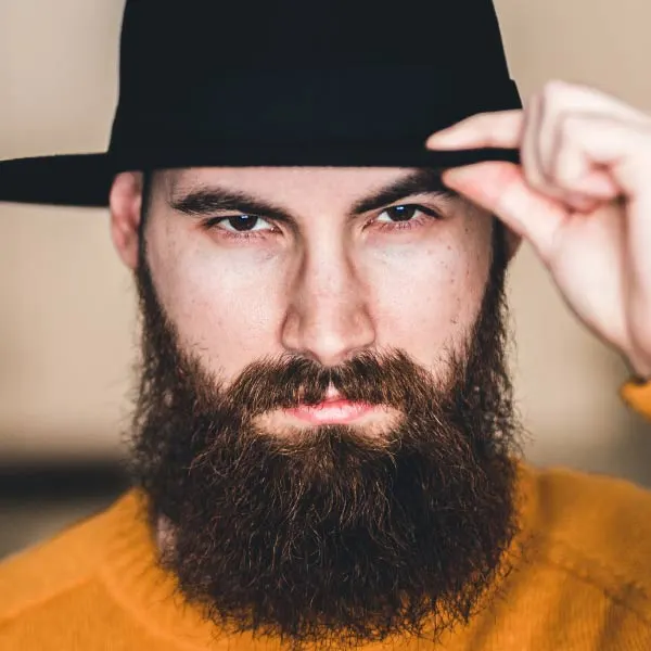 Man with beard wearing black hat and orange shirt, adjusting hat brim.