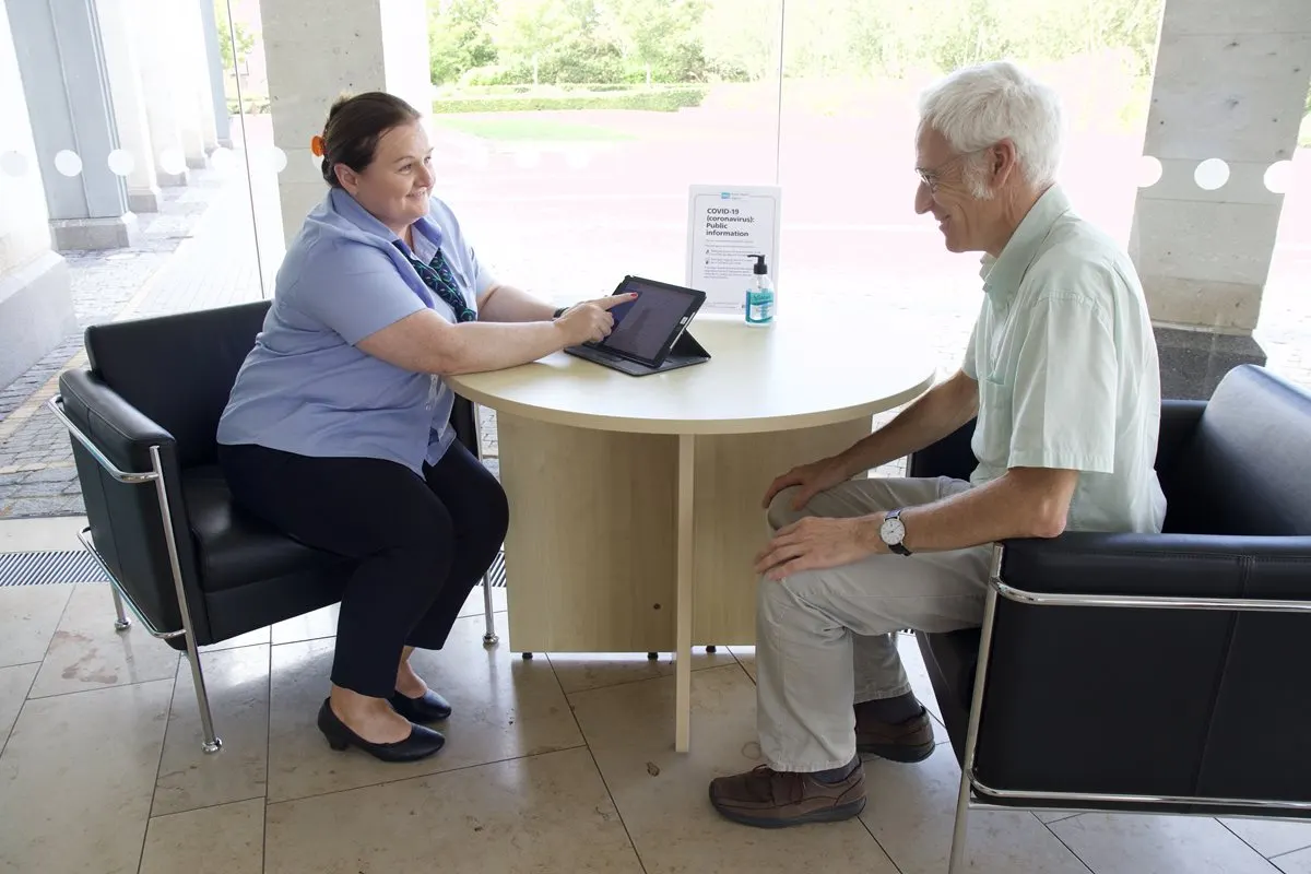 A staff member in a blue shirt holds a tablet whilst seated across from an older man at a round table in a bright, modern interior space.