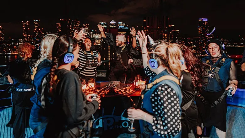 A group of people wearing headphones dancing and celebrating at night on a rooftop with a illuminated cityscape behind them.