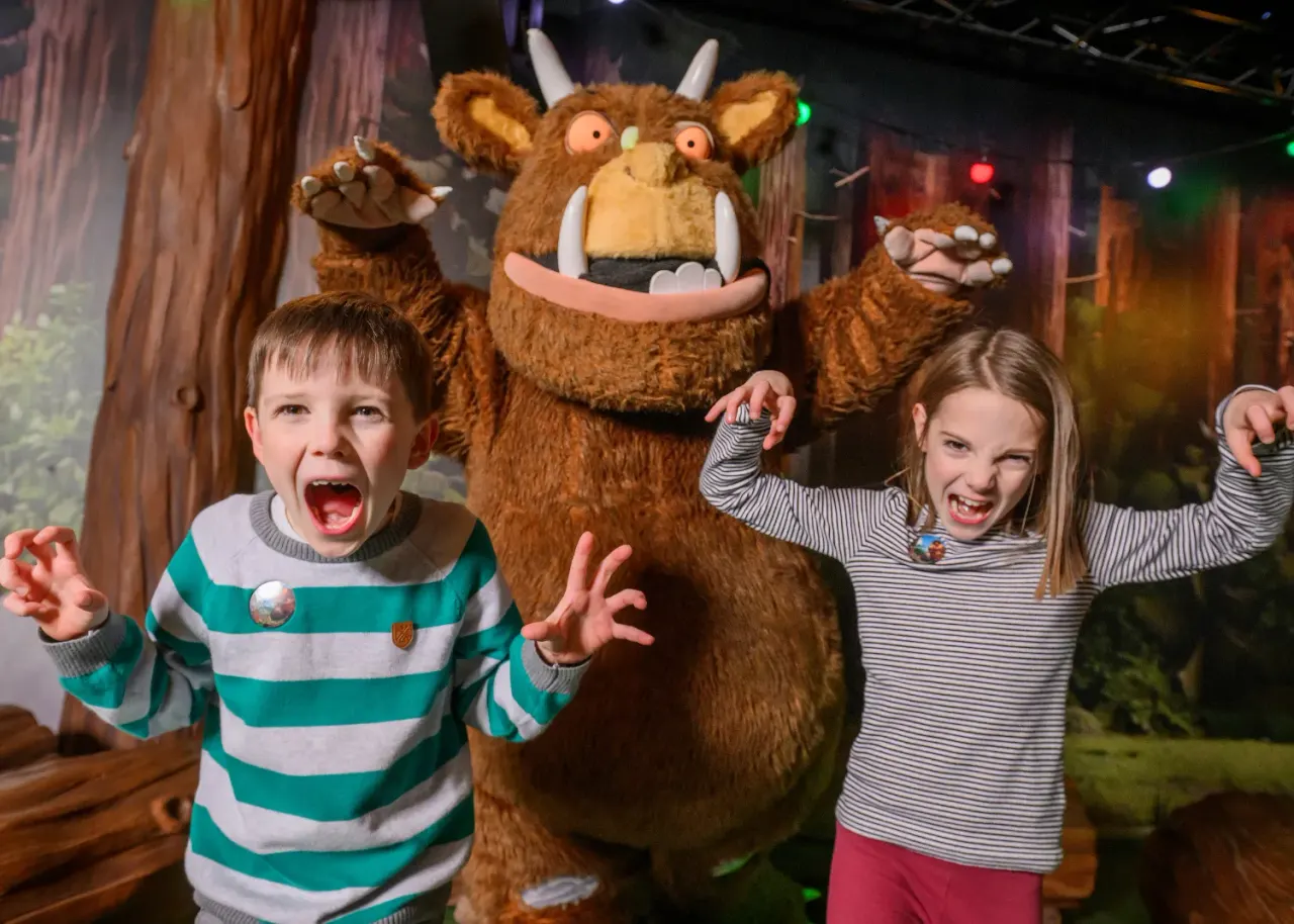 Two young children posing excitedly with a large Gruffalo character costume at an indoor attraction.
