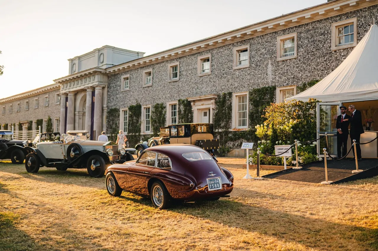 Classic car rally on the grounds of a grand stone mansion, with vintage vehicles displayed and visitors viewing them.