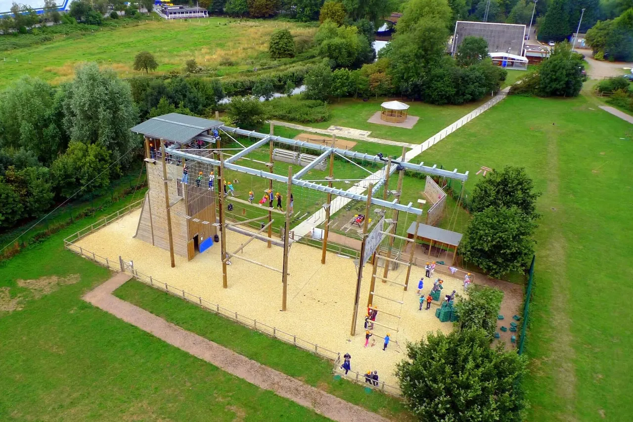 Aerial view of an adventure playground with wooden climbing structures, rope courses and climbing frames on sandy ground, surrounded by green lawns and trees.