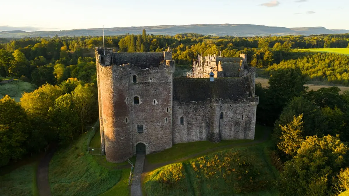 Medieval stone castle with towers surrounded by green grounds, forests and distant hills at sunset.