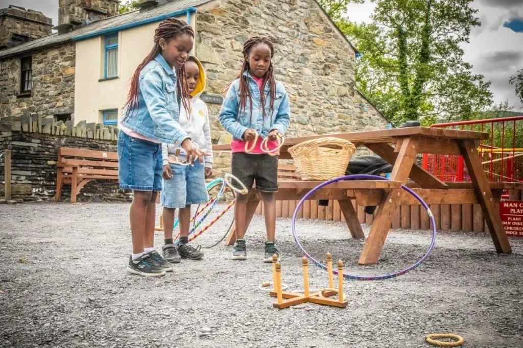 Three children play with hula hoops and quoits in a rural stone cottage garden setting.