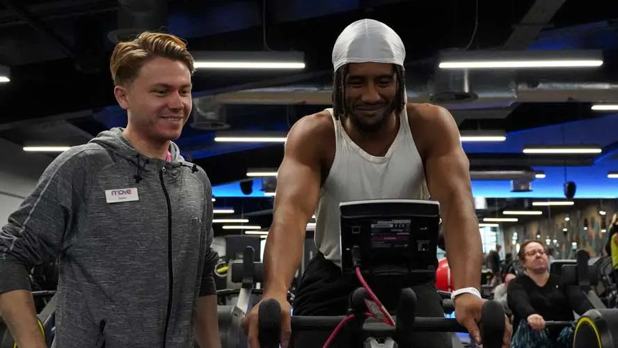 A man in a white cap and vest uses an exercise bike whilst a staff member smiles beside him in a modern gym.