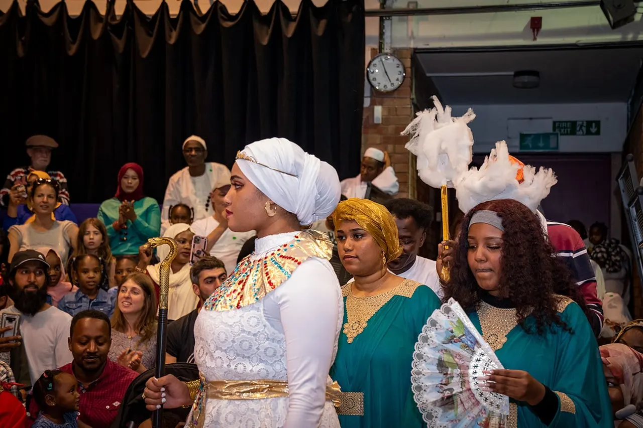 Performers in elaborate theatrical costumes with headpieces and fans perform on stage before an audience in an indoor theatre.