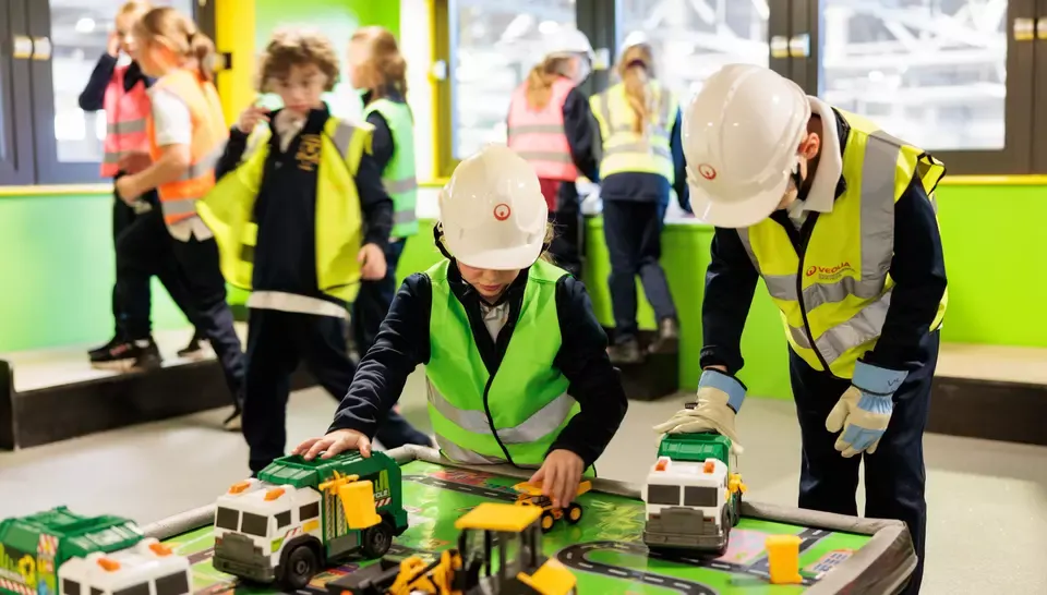 Children and adults wearing hard hats and high-visibility vests examine toy refuse vehicles at a recycling education workshop.