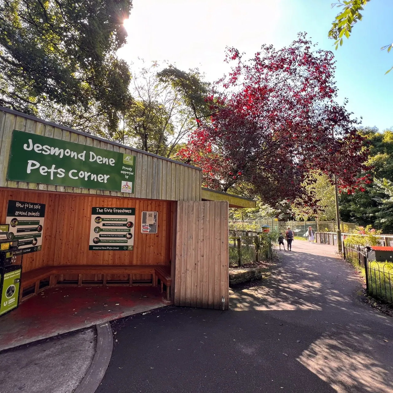 Entrance to Pets Corner with wooden shelter building, information boards, and visitors walking along tree-lined path in autumn.