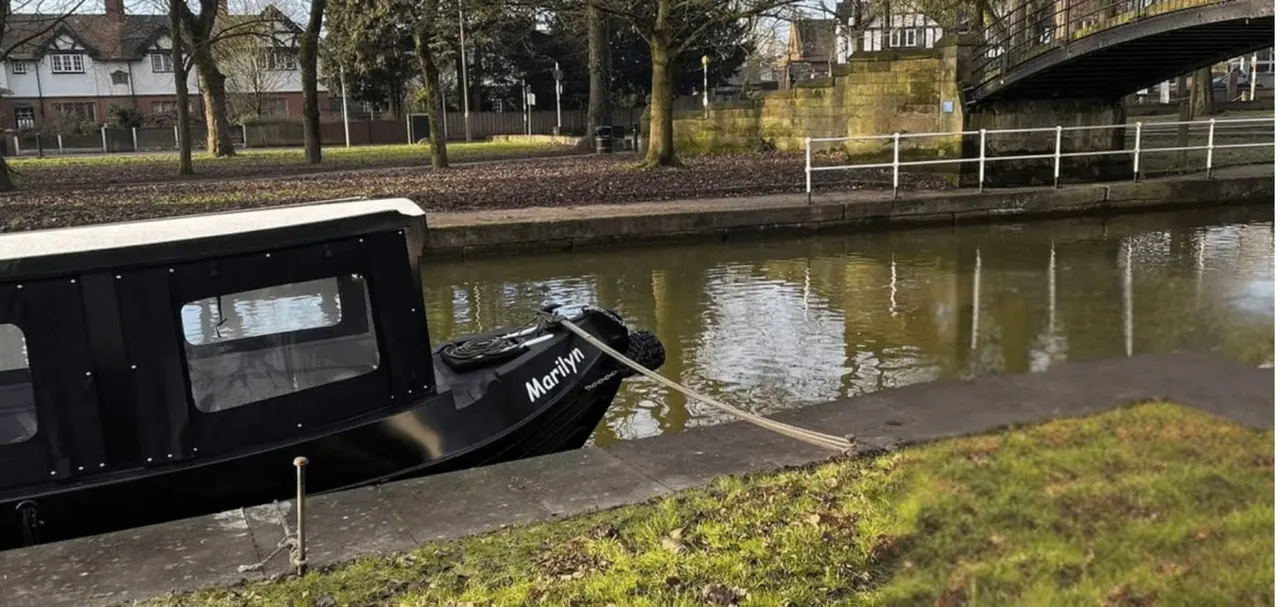 Black narrowboat moored on a canal beside a towpath, with urban buildings and a bridge visible in the background.