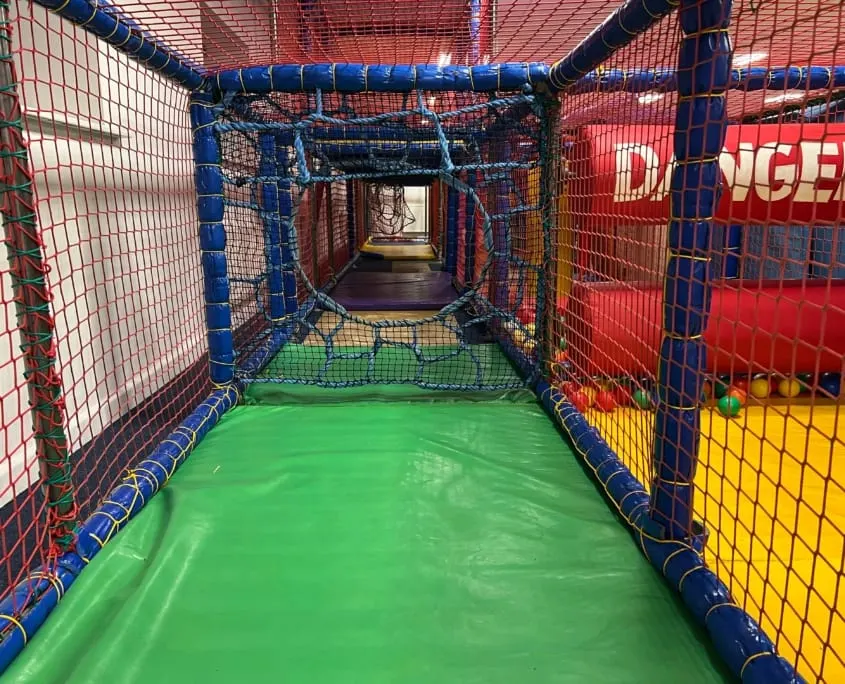 Interior view of a colourful soft play tunnel with green flooring and blue and red padded walls and netting.