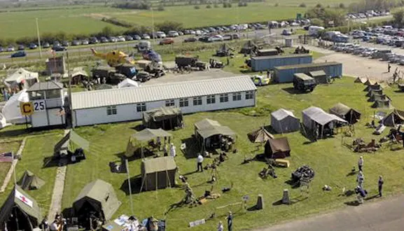 Aerial view of an outdoor event with visitors exploring military vehicles, tents, and equipment displays across a grassy field.