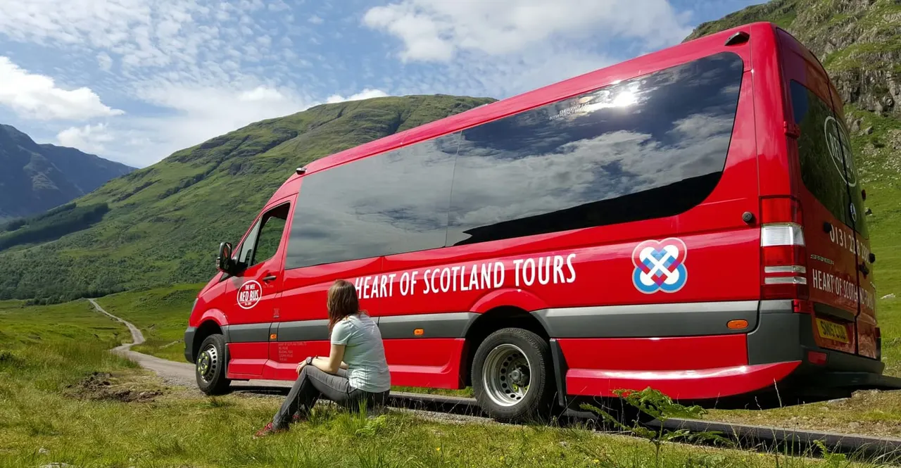 A person crouches beside a red tour bus in a remote Scottish valley with green hills and a winding road.