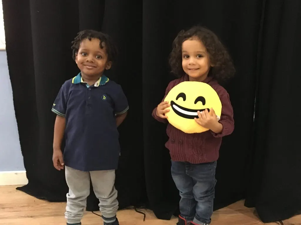Two young children smile at the camera against a black curtain backdrop, one holding a large yellow smiley face emoji prop.