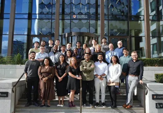 Large group of staff members posing outside a modern glass-fronted building in daylight.