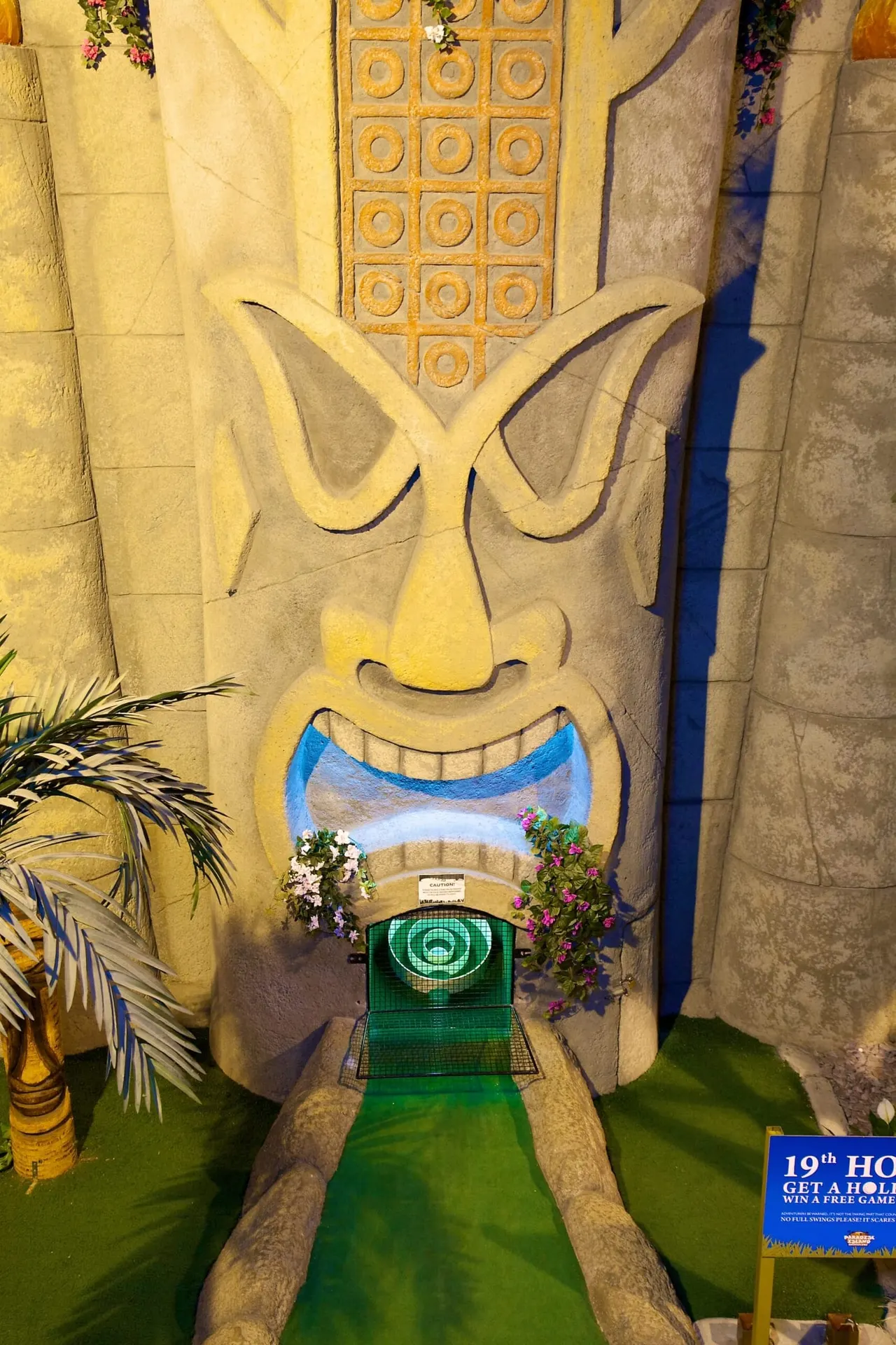 Miniature golf hole with bright yellow carved face sculpture and green putting surface.