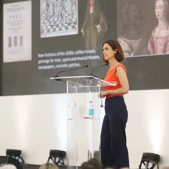 Woman in orange sleeveless top speaking at a lectern with a projected presentation about 18th-century coffee houses on the wall behind her.
