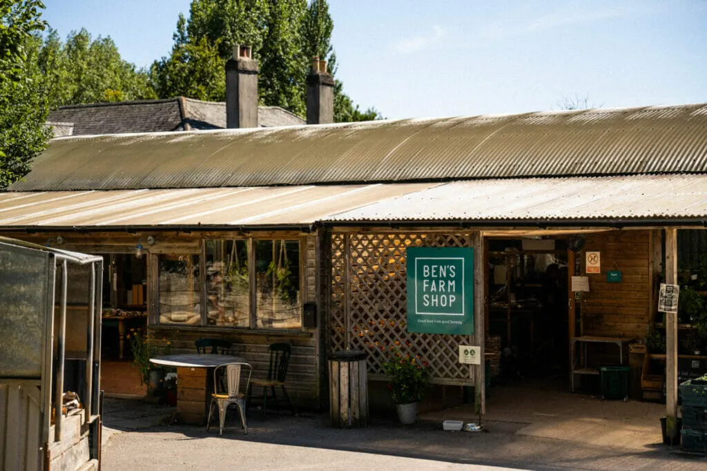 Rustic wooden farm shop building with corrugated metal roof, green signage, and outdoor seating area surrounded by tall trees.