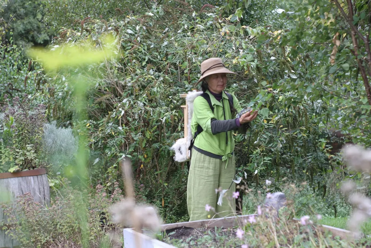 A woman in a green jacket and brown hat stands amongst overgrown plants and vegetation in a garden setting.