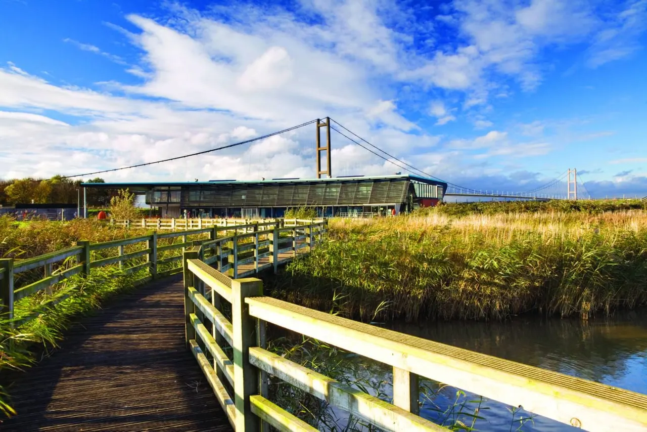 Wooden boardwalk and viewing platform overlooking wetland marshes with a modern visitor centre building and suspension bridge visible in the background.