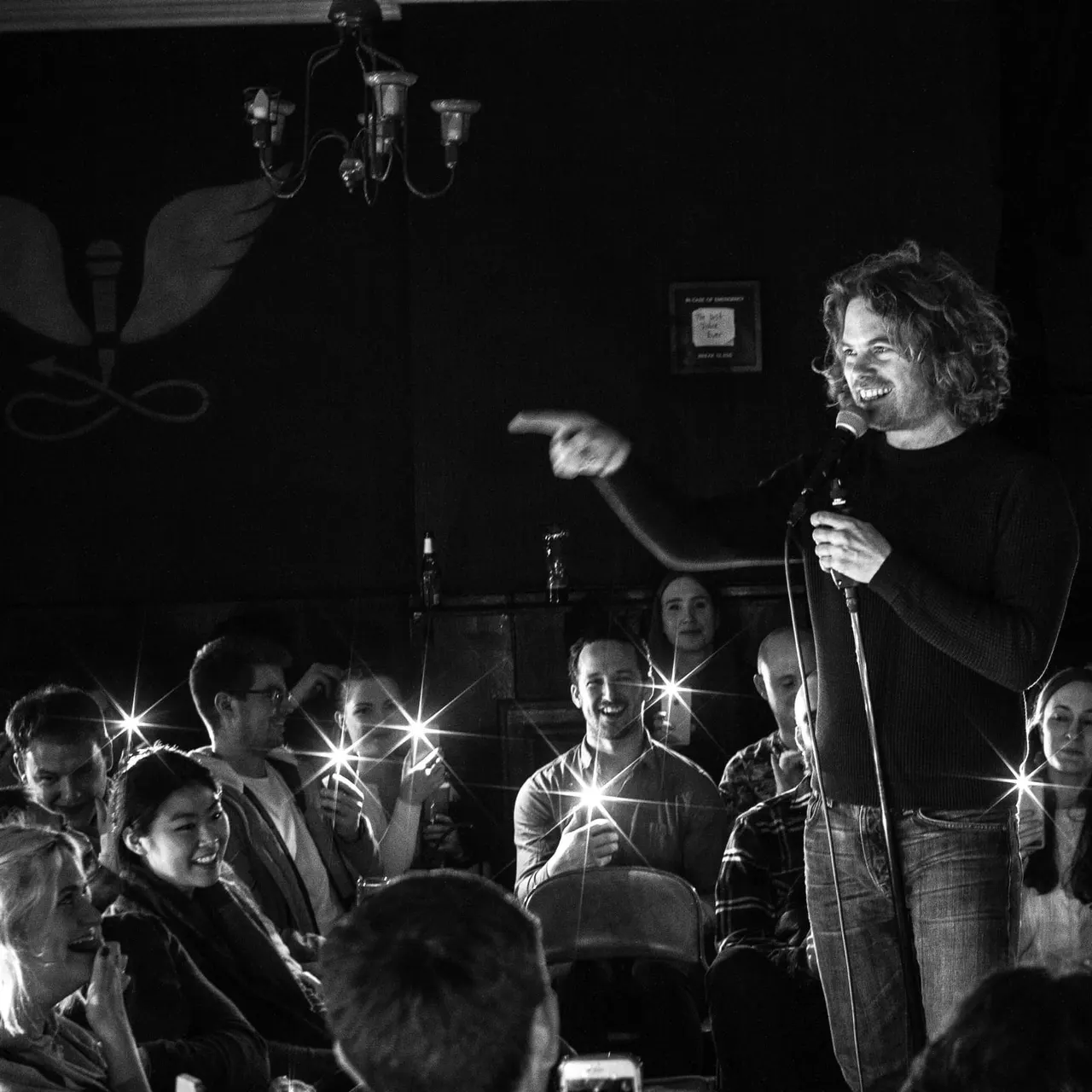 A performer holds a microphone on stage addressing a smiling audience in a dimly lit comedy venue.