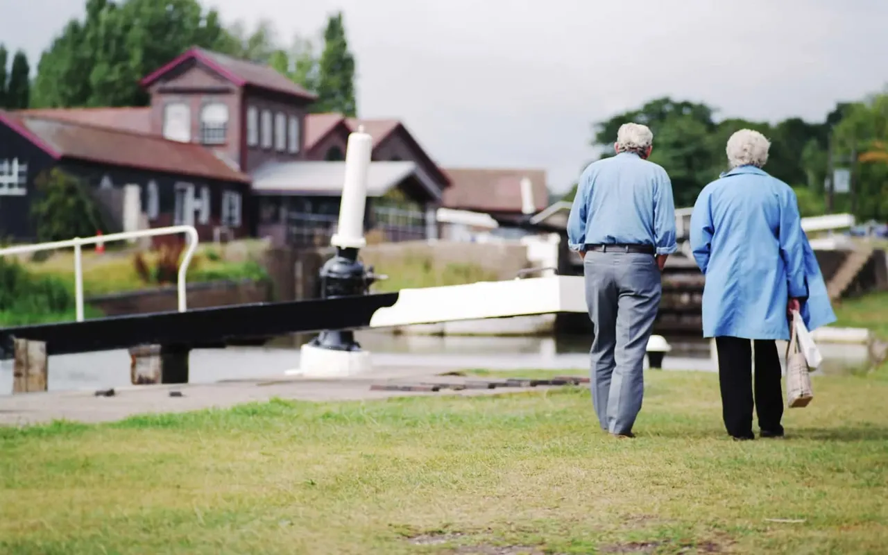 Two older visitors walk across a grass field towards canal lock gates and a red-roofed boathouse.