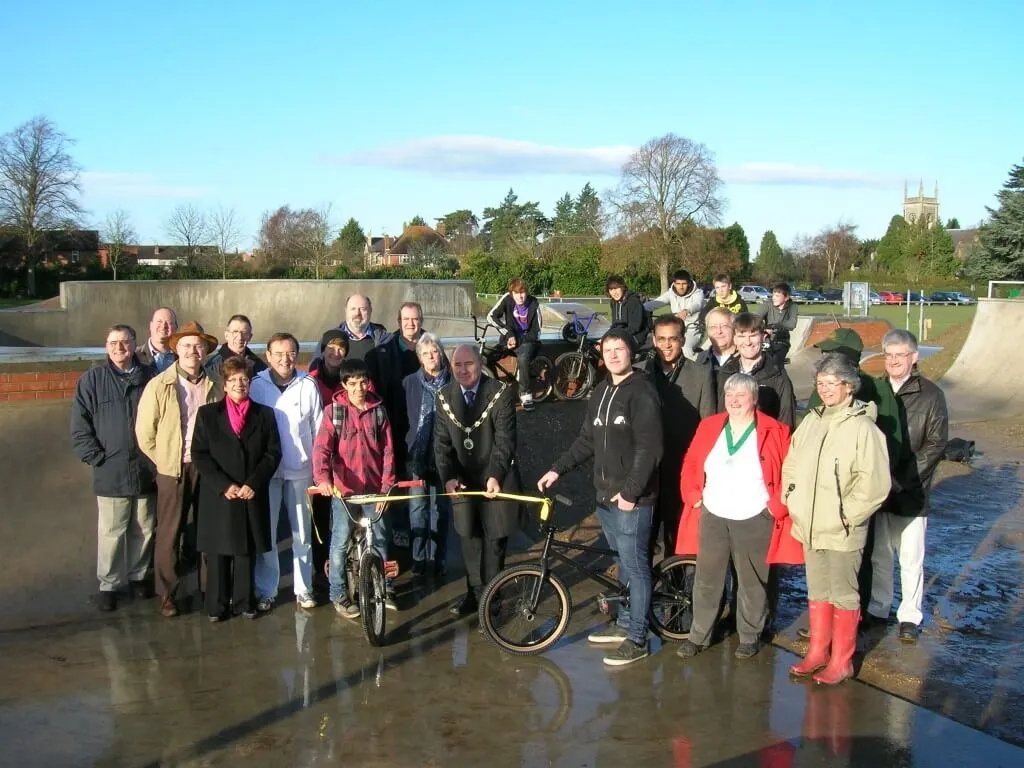 Large group of adults and young people gathered at a skate park with bicycles, posing for a photograph on wet ground.