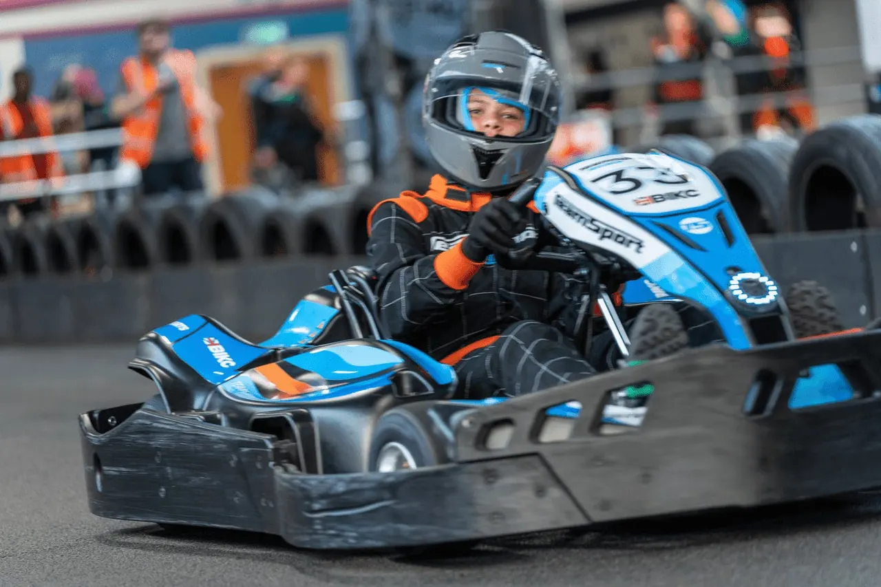 Child in helmet and orange safety gear driving a blue and white go-kart on indoor track with spectators behind barriers.