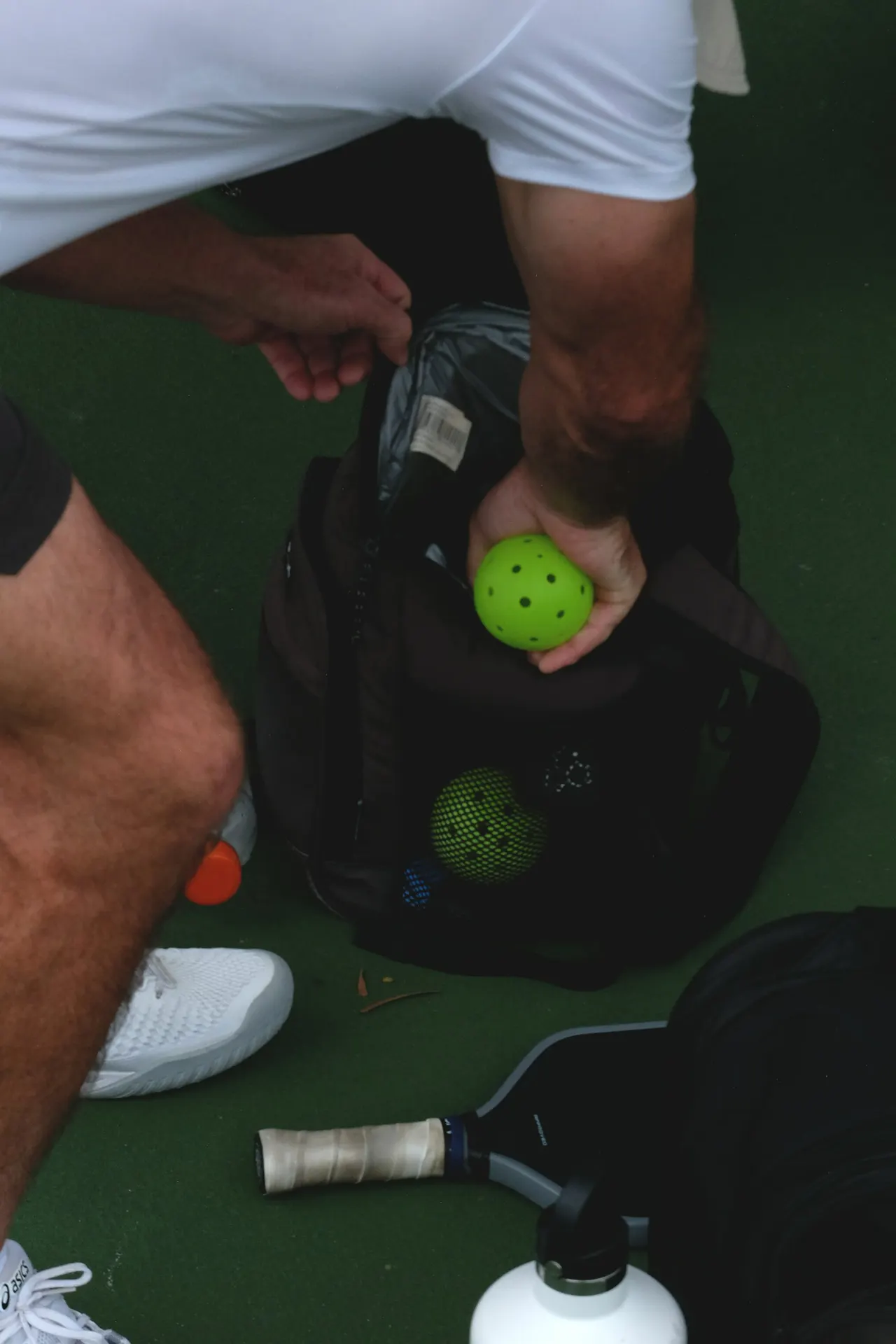 Person holding a bright yellow pickleball with holes, standing on a green court surrounded by paddles and equipment.