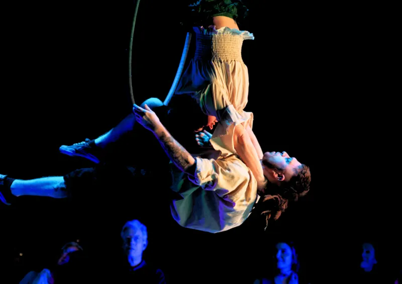 Acrobat performing an aerial trick suspended by rope above an audience in blue stage lighting.