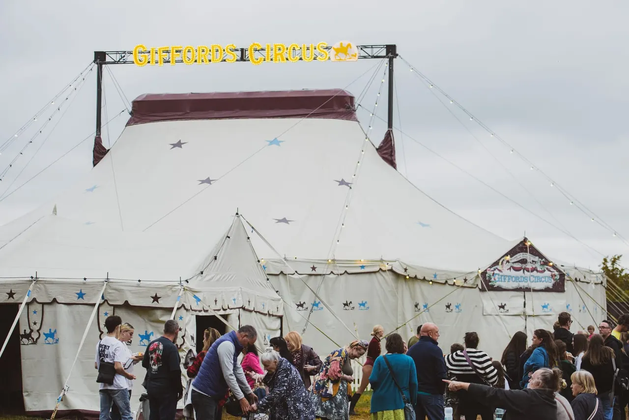 Large circus tent with cream canvas and burgundy trim, decorated with blue and grey stars, visitors queuing outside the entrance.