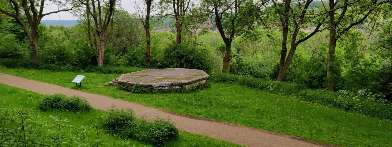 A curved stone platform set in a grassy woodland clearing with a path running alongside and countryside views beyond.