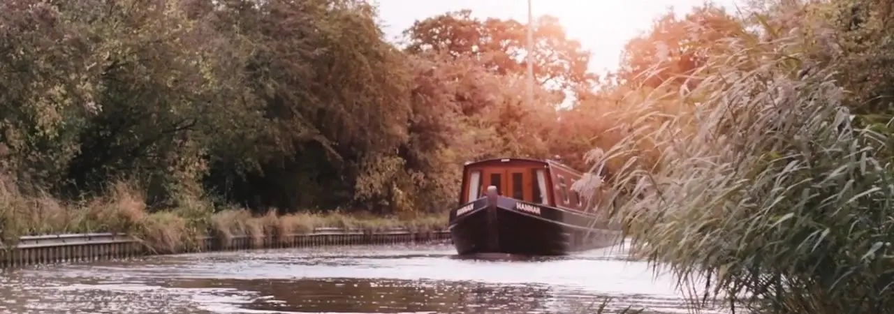 Narrowboat moored on a tree-lined waterway in autumn, surrounded by reeds and overhanging vegetation.