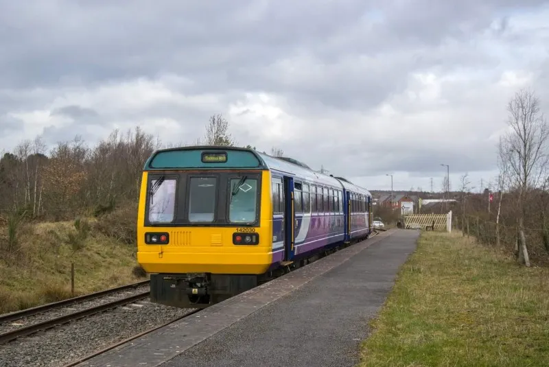 A purple and yellow passenger train stopped at a railway station platform with bare trees and countryside visible in the background.