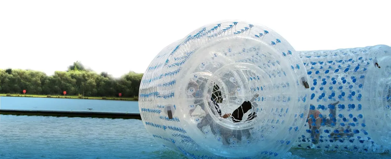 Child inside a large transparent inflatable water roller on a lake, with wooded shoreline behind.