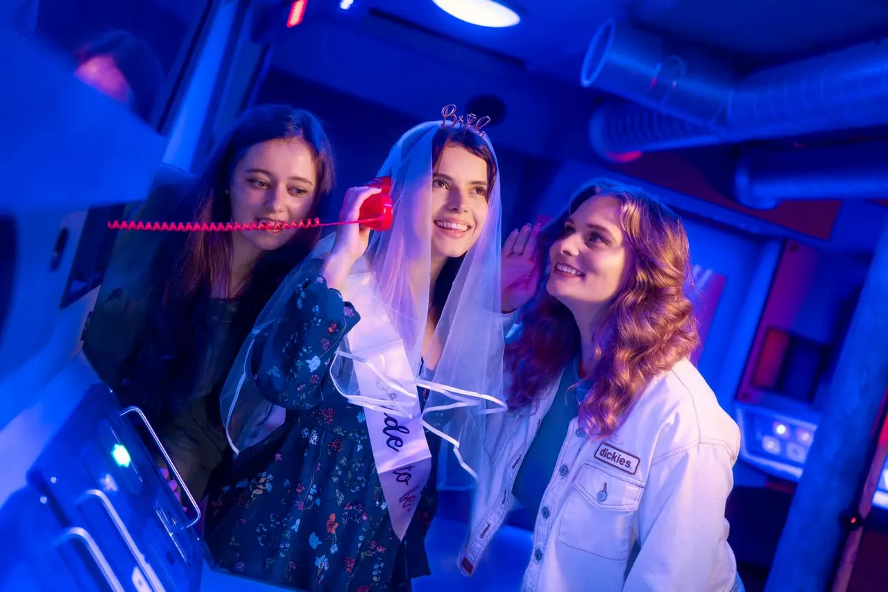 Three young women celebrating in a neon-lit room, with the centre woman wearing a white veil and "Bride" tiara.