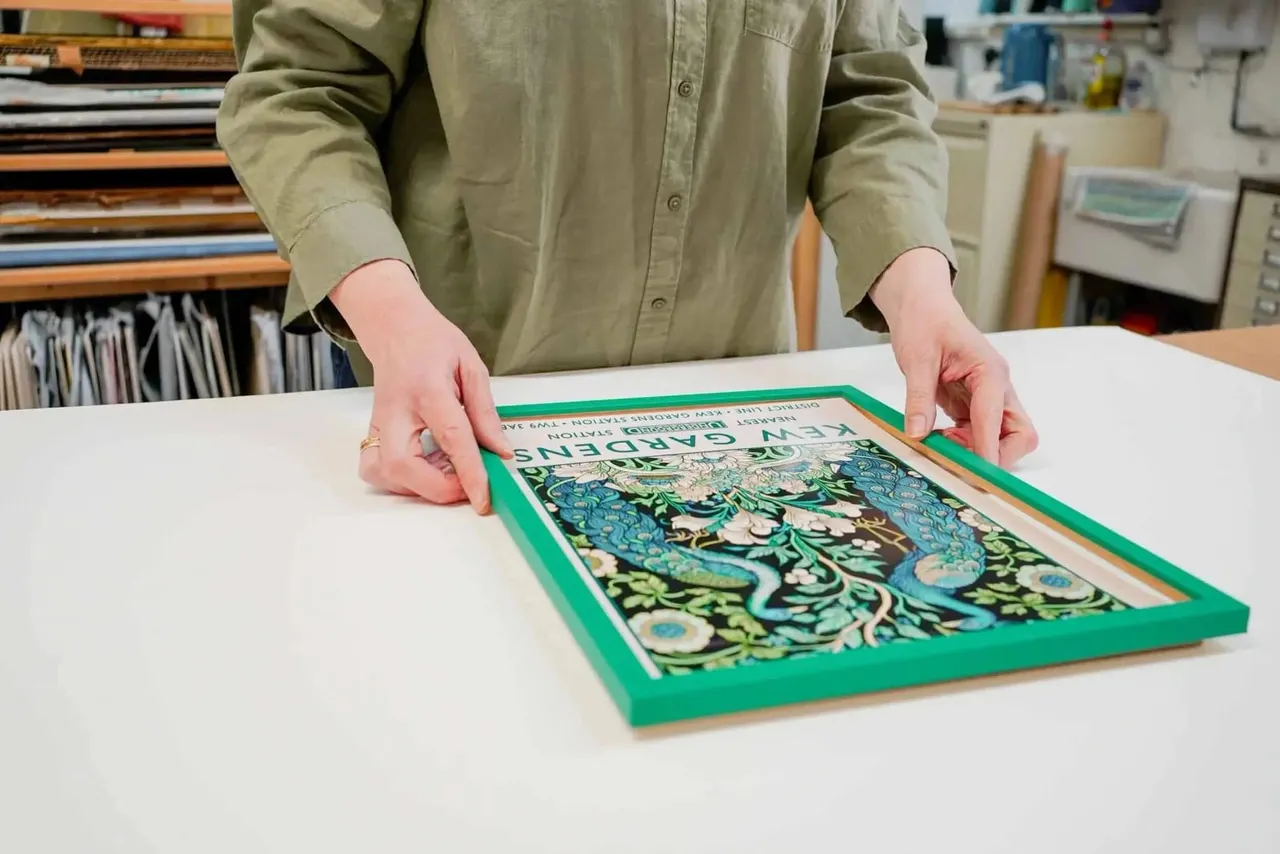 Person in khaki shirt displaying a framed botanical artwork with green border in a gallery workshop setting.