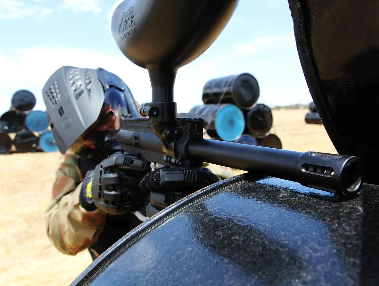 Close-up of paintball gun with hopper and scope on outdoor arena, players wearing masks and protective gear visible in background.