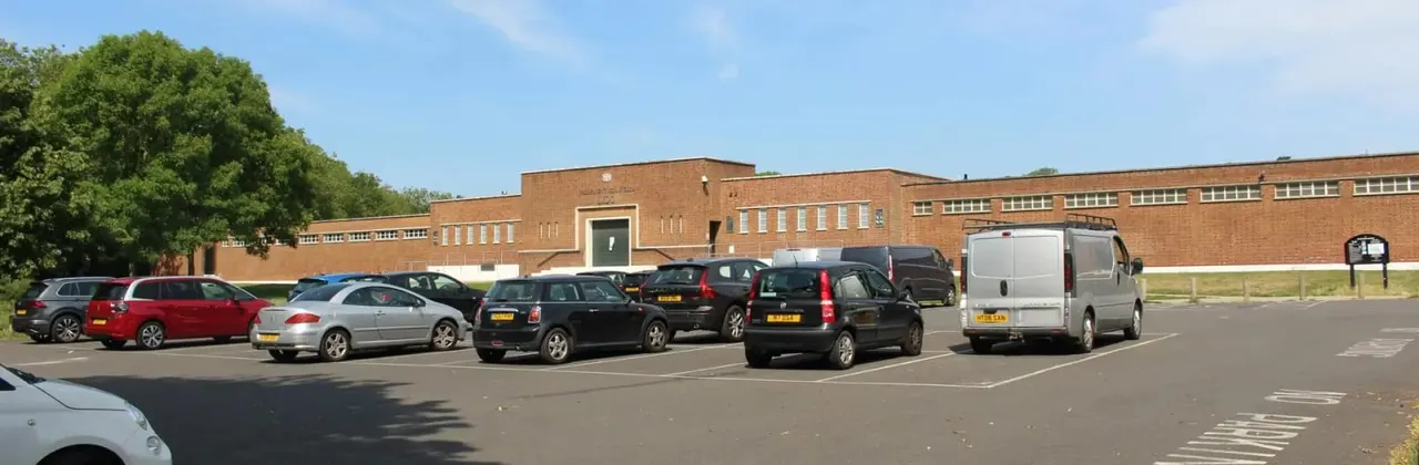 Cars parked in a tarmac car park beside a large brick building under blue sky.