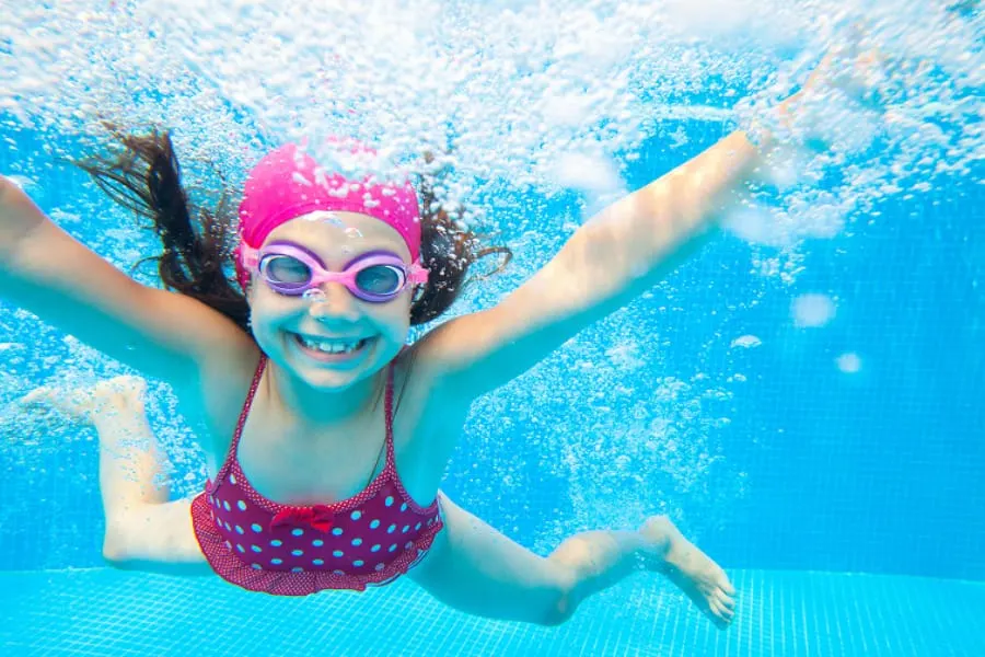 Child swimming underwater in pink swimwear and goggles, arms outstretched, smiling at camera.