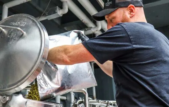 Man in cap pouring grain into brewery equipment during beer production.