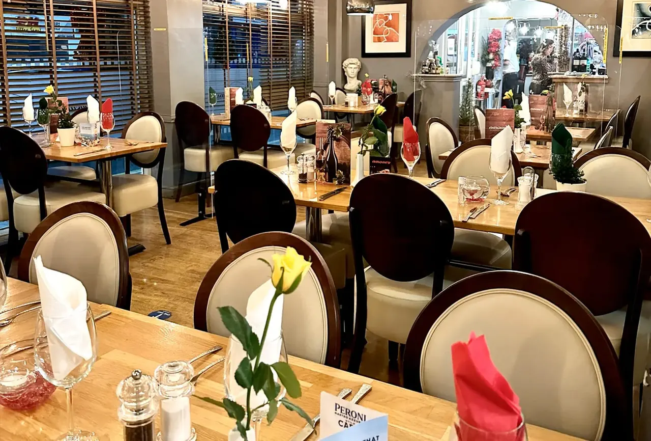 Elegant dining room with wooden tables set for service, upholstered chairs, fresh flowers, and modern décor with large windows overlooking the street.