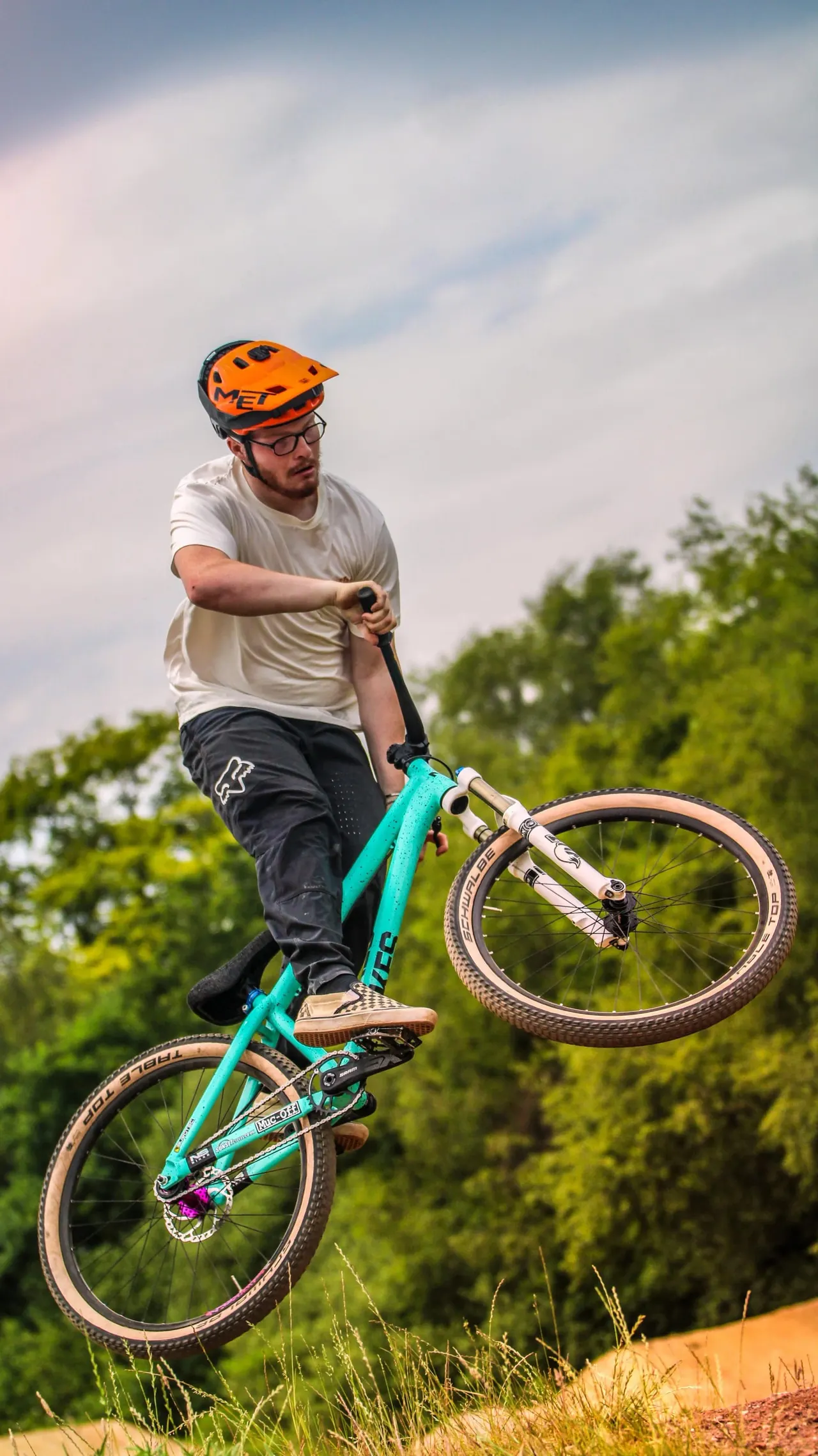 Man in orange helmet performing a jump on a turquoise mountain bike against a cloudy sky and green trees.