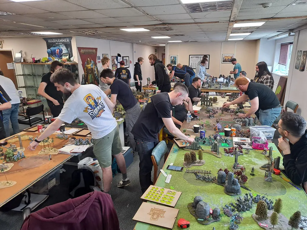 Players of various ages gather around tabletop gaming boards with painted miniatures and scenery in a community gaming space.