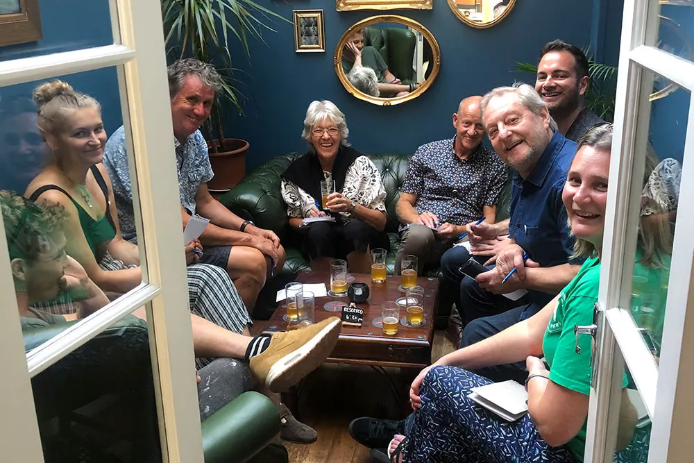 A group of eight adults socialising in a blue-walled room with drinks on a wooden table, surrounded by mirrors and potted plants.