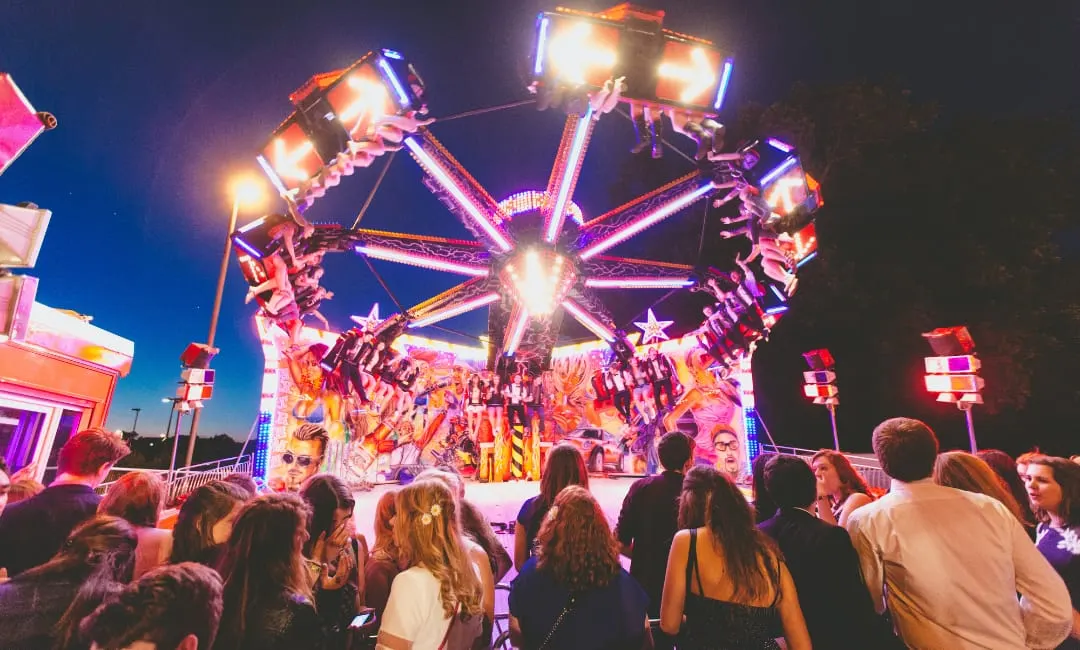 Crowd of people watching an illuminated fairground ride at night with bright pink and orange lights.