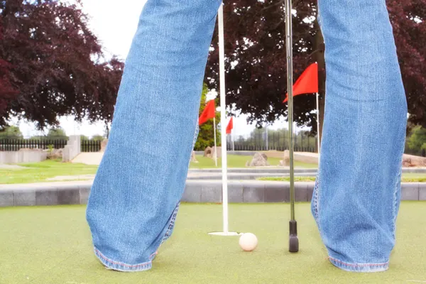 Person in jeans standing over golf ball near hole with flag on miniature golf course.