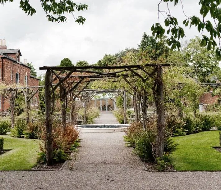 Pergola-covered pathway with climbing plants leading through manicured gardens towards a brick building.