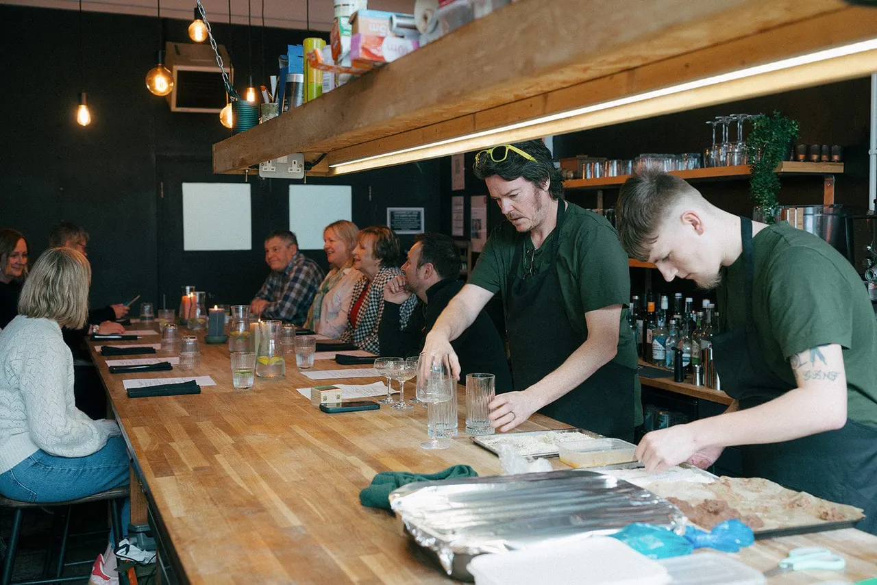 Staff in green shirts prepare a cooking demonstration at a wooden counter whilst guests watch seated along the bar in a modern kitchen venue.