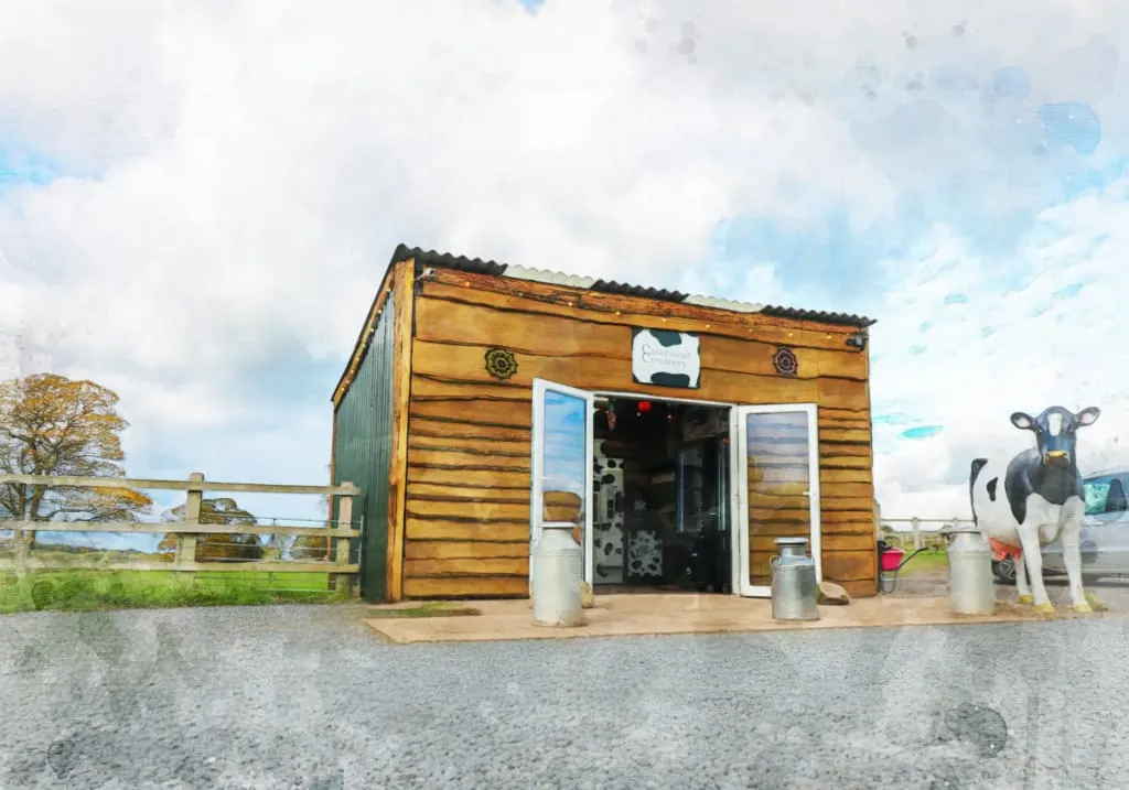 Wooden farm shop building with white doors and a cow statue outside, set in a rural paddock with fencing.